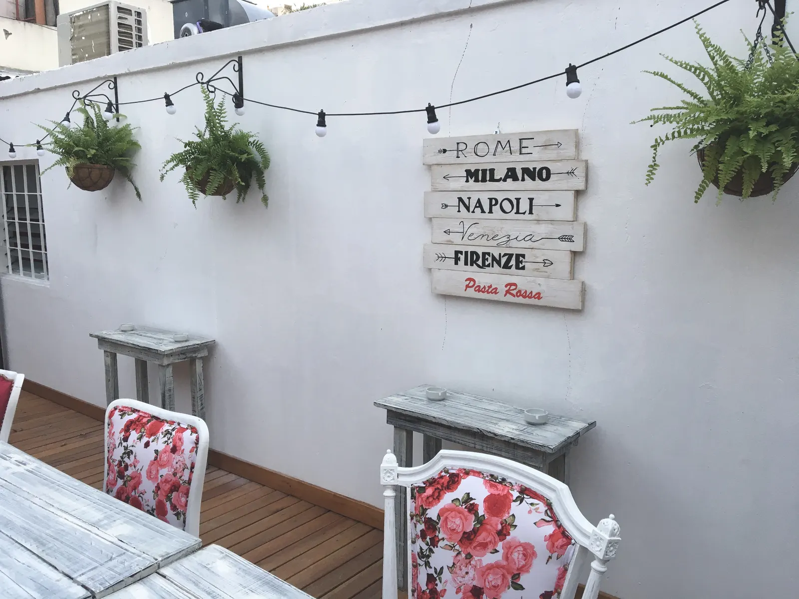 Terraza con plantas colgantes y cartel de ciudades italianas en Pasta Rossa Ramos Mejía